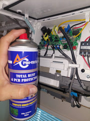 A hand holds a blue and white "AC Pro-Tech Total Rust & PCB Protection" spray can, aiming it at the intricate circuit board and wiring inside an open air conditioning unit. This demonstrates precise air conditioning maintenance, using a specialized pro tech cleaner to protect critical electrical components from corrosion.